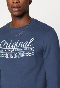 Navy plavi dukserica od pamuka, s bijelim izvezenim tekstom "Original CLAIM YOUR DENIM RIGHTS BLEND" i okruglim vratom. Vidljiv srebrni lanac.