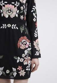 Robe noire à manches longues ornée de broderies en sequins multicolores représentant de grandes fleurs sur les manches, la taille et la jupe, portée par une personne avec la main détendue le long du corps.