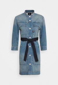 Jeansklänning i ljusblått med långa ärmar, knappstängning fram, två bröstfickor och midjesnörning. Har Vita knappar och ett kontrasterande svart bälte.