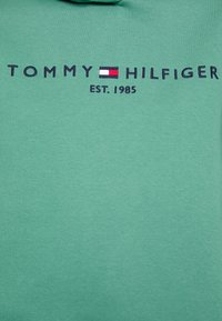Logo Tommy Hilfiger s červenou, bílou a modrou vlajkou a nápisem "EST. 1985" vyšitým na zeleném textilním pozadí.