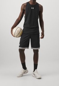Débardeur de sport noir et short avec bordure blanche, tenant un ballon de basket. Le maillot porte le logo Under Armour sur la poitrine. Baskets grises.