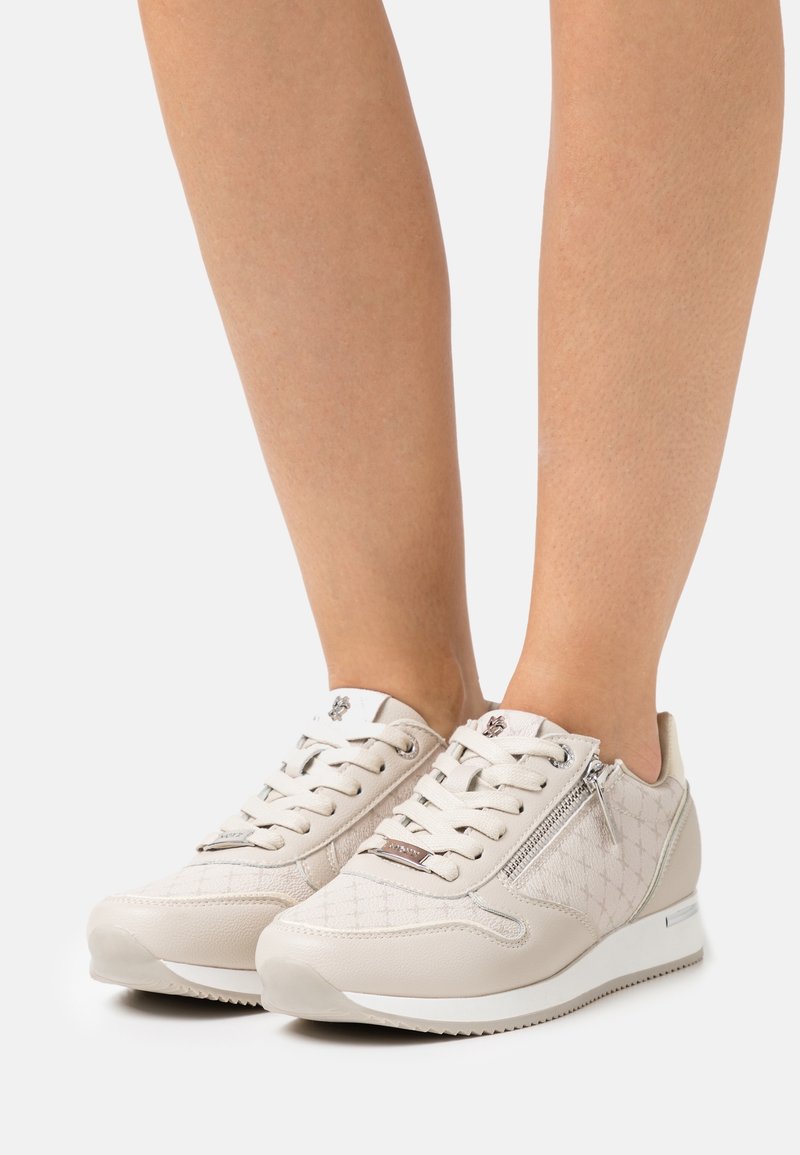 Baskets basses blanches avec motif matelassé et fermeture éclair latérale portées sur des jambes nues sur un fond uni.