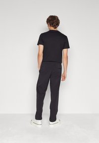 Sort tætsiddende t-shirt og løse sorte bukser, begge lavet af blødt stof. Modellen har hvide sneakers med en sort streg på, set bagfra.