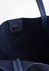 Tmavě modrá taška vyrobená z semiše, s hladkým vnitřním podšívem a kulatým kovovým zacvakáváním na vrchu.