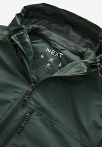 Veste imperméable vert foncé doublée en polaire avec capuche, doublure en mesh, deux poches zippées, et étiquette de la marque NEXT indiquant la taille moyenne.