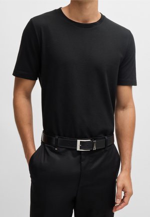 Mann trägt ein eng anliegendes schwarzes, strukturiertes Kurzarmhemd, schwarzen Gürtel mit rechteckiger silberner Schnalle und schwarze Hose, Hände in den Taschen.