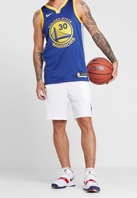 Uomo che indossa una maglia da basket dei Golden State Warriors, tiene un pallone da basket, indossa pantaloni corti bianchi e scarpe sportive, in piedi contro uno sfondo neutro.