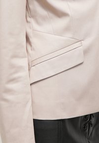 Détail de rabat de poche de veste beige sur pantalon en cuir noir avec un laçage visible sur le côté.