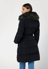 Parka nera con ampio colletto in pelliccia sintetica, texture trapuntata, vita con cintura e maniche lunghe, indossata sopra jeans blu.