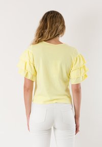 Top giallo a maniche corte con maniche sovrapposte e a volant. Materiale in cotone morbido. Orlo leggermente curvo, indossato con pantaloni bianchi.