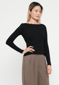 Camisa de manga larga negra con textura acanalada y borde de encaje con volantes alrededor del escote, combinada con pantalones plisados de color marrón claro.