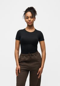 Γυναίκα με σγουρά μαλλιά σε κότσο που φοράει εφαρμοστό μαύρο μπλουζάκι και καφέ φόρμα με κορδόνι, στέκεται μπροστά σε λευκό φόντο.