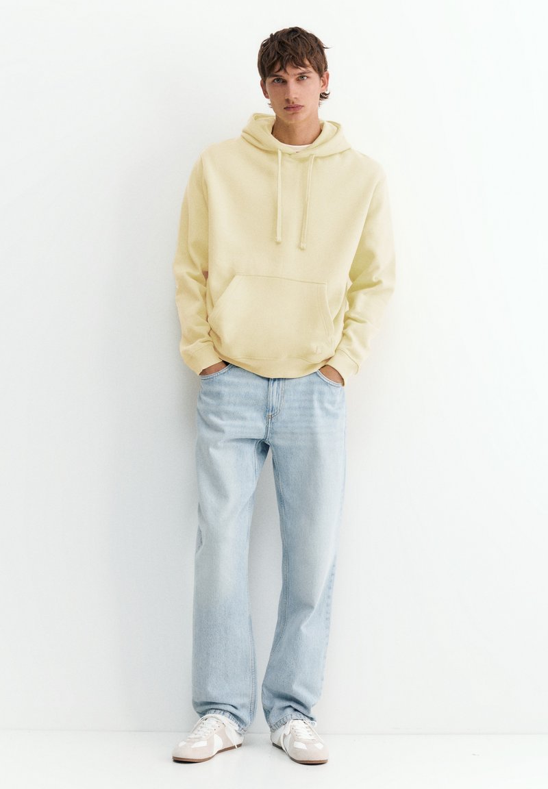 Jeune homme debout, les mains dans les poches, portant un sweat à capuche jaune clair, un jean bleu clair et des baskets blanches sur fond blanc.