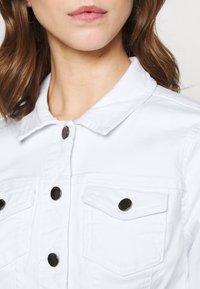 Mujer vestida con una chaqueta de denim blanca con botones negros y un cuello, mostrando el cuello y parte del cabello castaño.