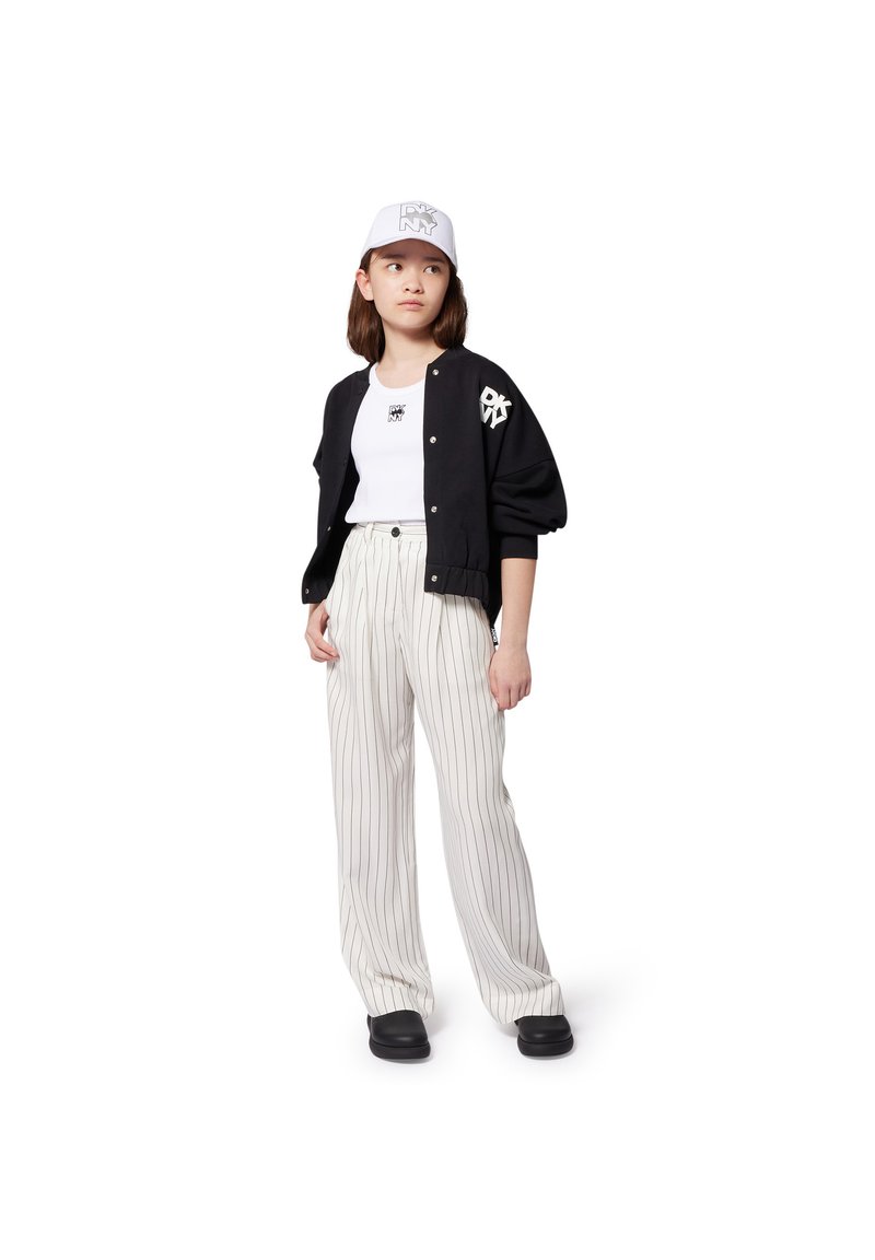 Giubbotto bomber nero con logo DKNY, t-shirt bianca, pantaloni a palazzo beige a righe, e cappellino bianco con logo DKNY e visiera anteriore. Scarpe nere.