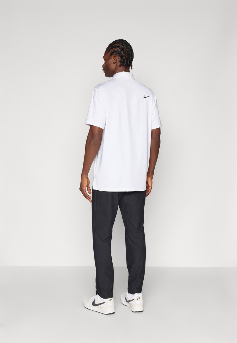 Polo bianco con logo Nike nero sulla schiena, abbinato a pantaloni scuri e scarpe da ginnastica chiare, visto da dietro.