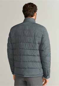 Chaqueta gris acolchada con cuello alto, costuras horizontales y una textura suave. Cuenta con un botón en el puño.