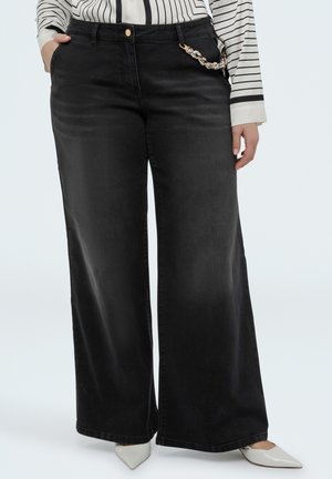 Donna con jeans neri a zampa con catena sulla tasca, maglia a righe a manica lunga e scarpe bianche con punta a becco, in piedi con una mano in tasca.