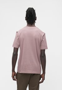 Bărbat cu dreadlocks, purtând un tricou roz prăfuit și pantaloni maro, stând cu spatele către cameră, pe un fundal deschis, simplu.