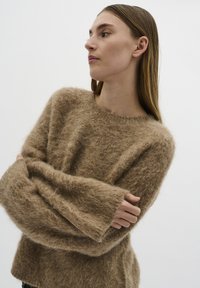 Brązowy, puszysty sweter z dzianiny o luźnym kroju, szerokimi rękawami i okrągłym dekoltem. Tekstura wydaje się miękka i puszysta, co zwiększa komfort cieplny.