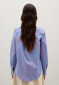 Camisa de manga larga de algodón en azul claro con rayas verticales rojas y blancas, con puños abotonados y un dobladillo curvado, que muestra un ajuste relajado.