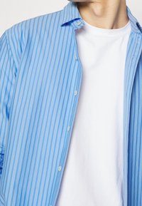 Hellblaue Button-up-Bluse mit vertikalen weißen Streifen, kragenloses Design und einem weißen Rundhalsausschnitt-T-Shirt darunter.