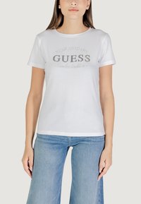 T-shirt bianco in cotone con logo argentato "GUESS" accompagnato da "ESTABLISHED 1981" e il testo "Tradizione senza tempo", colletto rotondo e maniche corte.