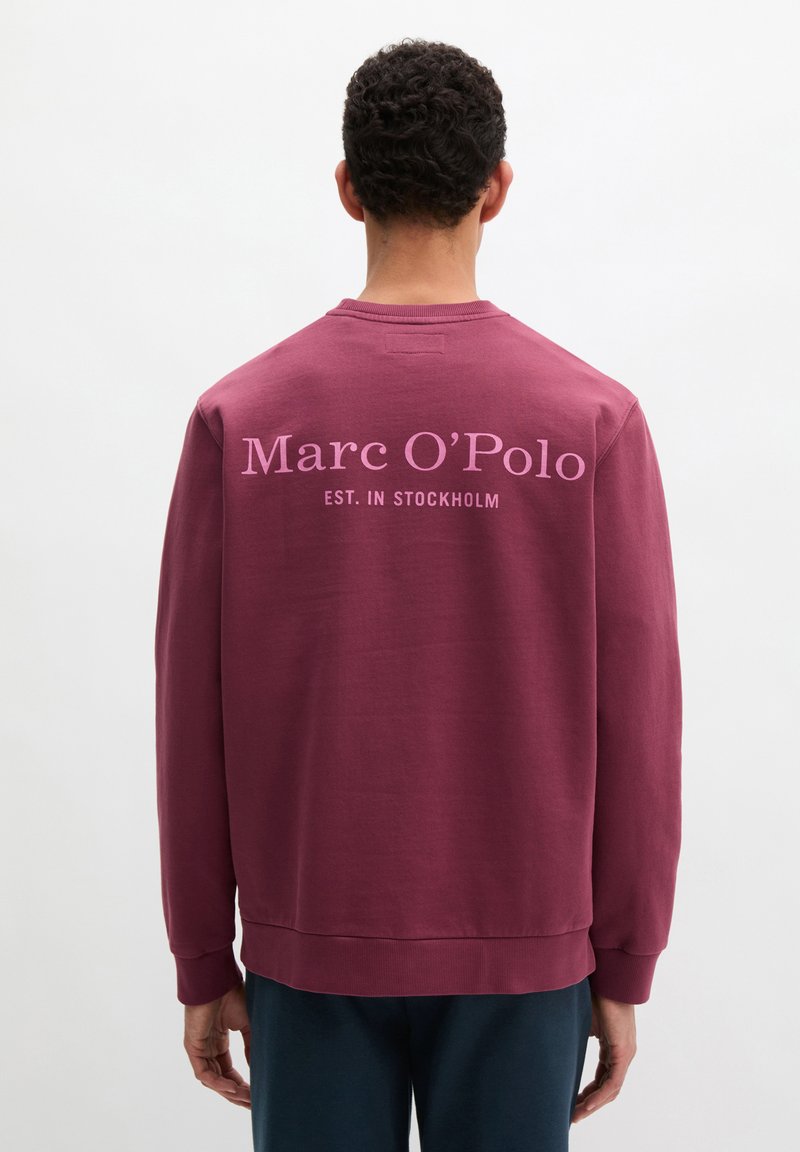 Άνδρας που φοράει μπορντό φούτερ με την επιγραφή "Marc O’Polo EST. IN STOCKHOLM" τυπωμένη με ροζ στο πίσω μέρος, στέκεται μπροστά σε απλό φόντο.