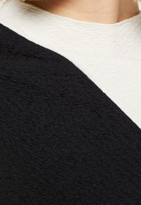 Schwarzer, strukturierter Stoff mit einem gekräuselten Muster kollidiert mit glattem Elfenbein-Stoff und präsentiert ein asymmetrisches Design am Ausschnitt.