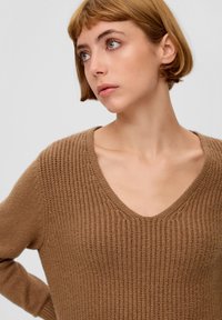 Pull en tricot marron avec un design en V, présentant une texture côtelée et des manches longues. Le matériau semble doux et confortable.