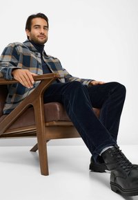 Geruite flanellen jas in marineblauw, grijs en beige, gecombineerd met donkere corduroy broek en zwarte veterschoenen, zittend in een houten stoel.