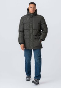 Mørkegrøn pufferjakke med hætte, der har et quiltet design, trykknaplukninger og frontlommer, båret over blå jeans og grå sneakers.