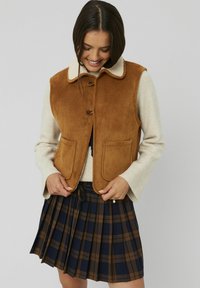 Veste en daim marron avec un col en fourrure crème, dotée de grandes poches plaquées. Portée sur un pull beige et une jupe plissée à carreaux bleu marine et marron.