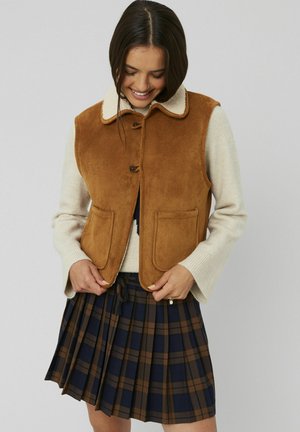 Veste en daim marron avec un col en fourrure crème, dotée de grandes poches plaquées. Portée sur un pull beige et une jupe plissée à carreaux bleu marine et marron.