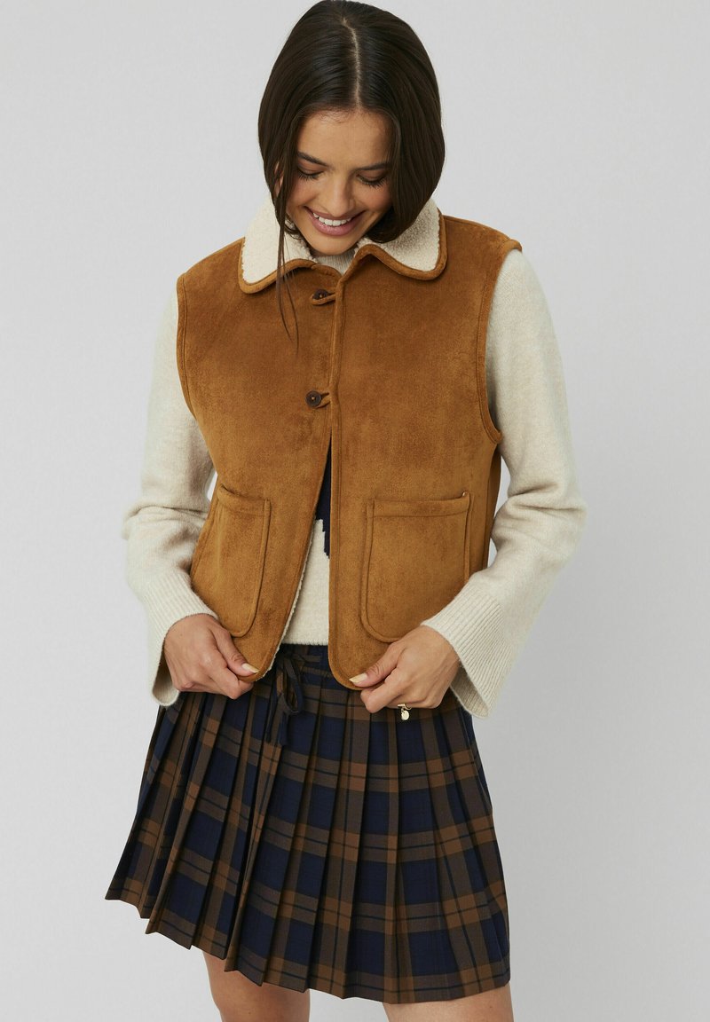 Veste en daim marron avec un col en fourrure crème, dotée de grandes poches plaquées. Portée sur un pull beige et une jupe plissée à carreaux bleu marine et marron.