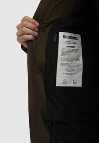 Chaqueta exterior verde oliva con un interior negro. Presenta un bolsillo con cremallera y una etiqueta blanca con texto. Incluye un cierre de botón a presión.