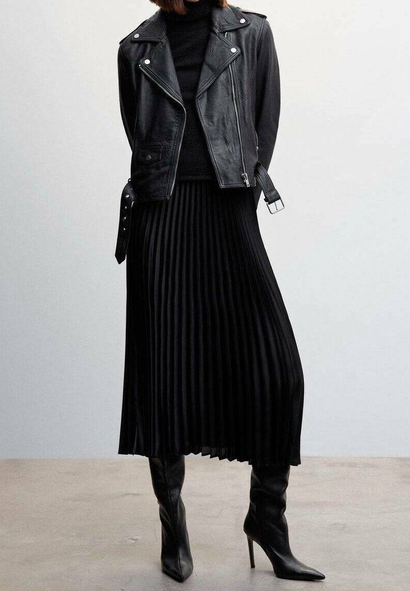 Personne portant une veste de motard en cuir noir, une jupe midi plissée noire, un col roulé noir et des bottes à talons hauts noires, debout à l'intérieur.
