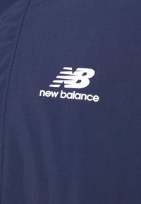 Tmavě modrá tkanina s hladkou texturou; obsahuje bílý logotyp New Balance v moderním písmu s elegantním designem.