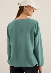 Losse, lange mouwen shirt van teal stof met een brede halslijn en subtiele glans, gecombineerd met een witte baseballcap.