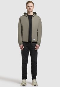 Olivgrüne Zip-Jacke mit Kapuze, elastischen Bündchen und einer Fronttasche, kombiniert mit einem schwarzen Shirt und dunklen Jeans.