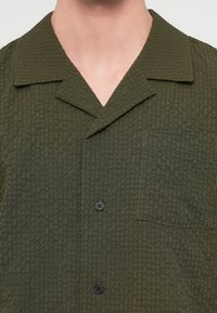 Κοντινό πλάνο ενός υφασμάτινου σκούρου πράσινου πουκάμισου με γιακά και τσέπη στο στήθος σε ένα άτομο.