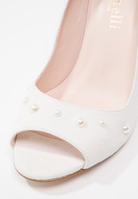 Escarpin en daim blanc à talon haut avec un bout ouvert, présentant un intérieur lisse rose clair et des embellissements en perles blanches le long de la sangle.