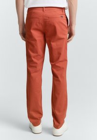 Un pantalon de couleur rouille en coton, avec une coupe droite, deux poches arrière et des baskets blanches. Texture lisse, coupe décontractée.
