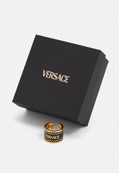 Anillo de metal dorado con una banda negra, que presenta "VERSACE" en letras doradas y un patrón de tablero de ajedrez. Incluye una caja negra con el logotipo.