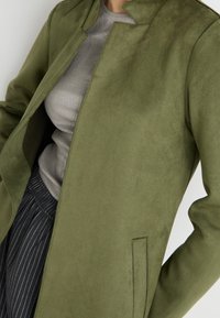 Chaqueta de ante sintético verde oliva con cuello alto, apertura frontal y bolsillos laterales, llevada sobre una blusa gris de canalé y pantalones a rayas.