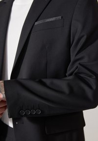 Sacou negru cu un design elegant, un singur buzunar pe piept cu un accent cu model și detalii cu patru nasturi pe manșetă. Textură de material neted.