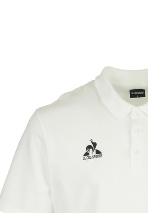 Polo blanc avec un col, présentant un logo noir sur la poitrine gauche. Il dispose d'un plastron à boutons et de manches courtes.