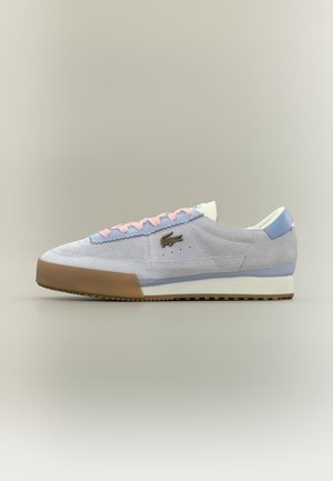 Gaiši pelēks zamšādas sporta apavs ar pasteļvioletiem un rozā akcentiem, brūnu gumijas pirkstu aizsargu, baltu zolīti un tumši zaļu krokodila izšūtu logotipu.