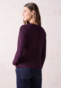 Lila geripptes Longsleeve-Oberteil mit Rundhalsausschnitt, ausgestattet mit seitlichen Schlitzen. Kombiniert mit dunkelblauen Denim-Hosen, die Taschen auf der Rückseite zeigen.