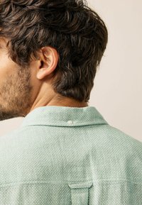 Green textured shirt with a collared neckline, featuring a single pearl button at the back. Fabric appears soft and lightweight.